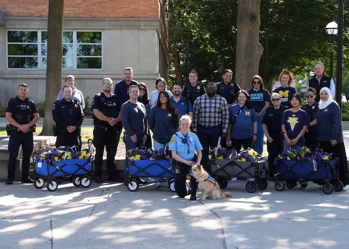 Police officers and volunteers including Wolverine Wellness staff pose with three blue wagons filled with University of Michigan merchandise and a police service dog during a community event.