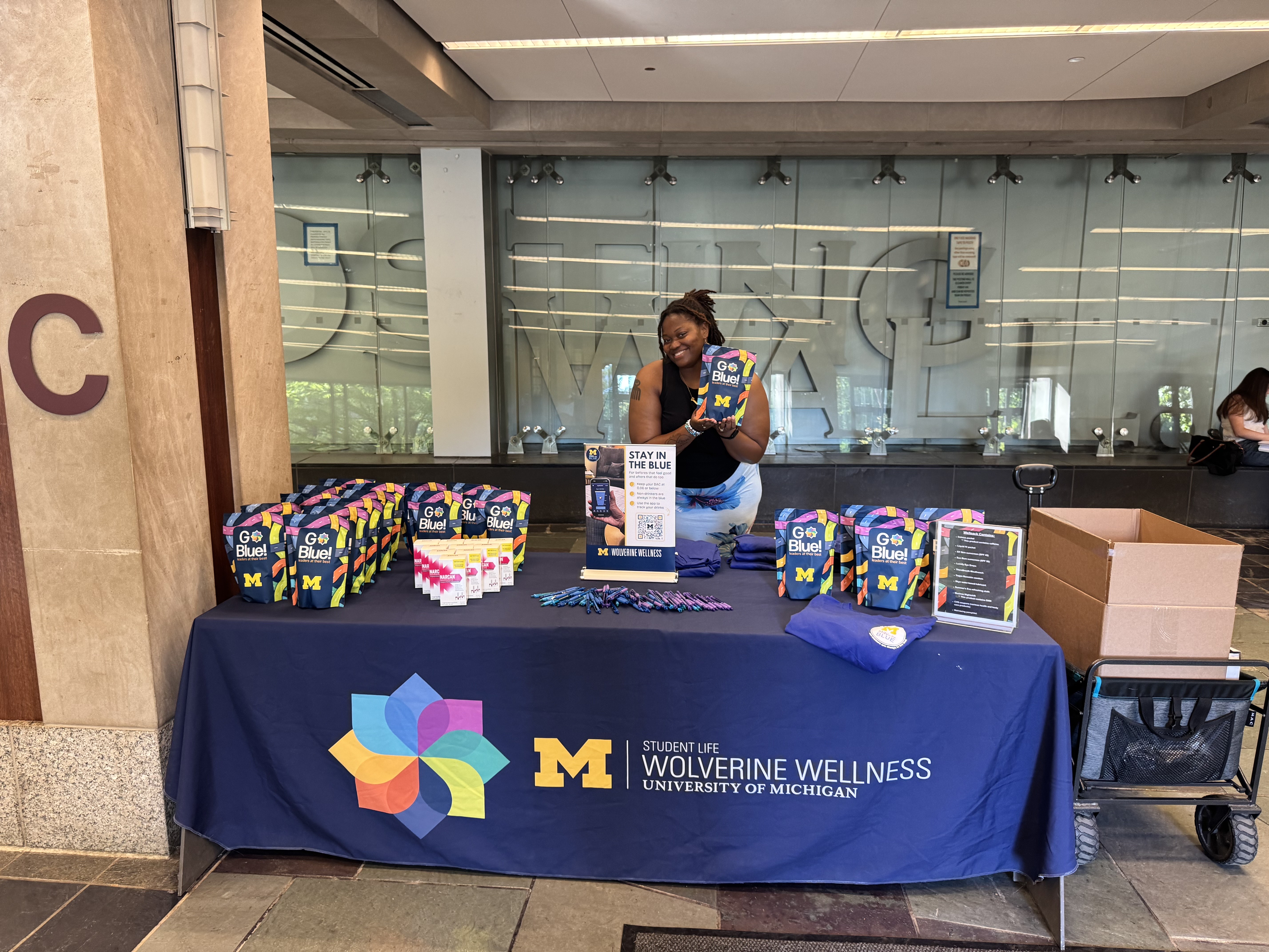 Student staff member at Wolverine Wellness table holding Go Blue wellness pack at Stay in the Blue popup event.