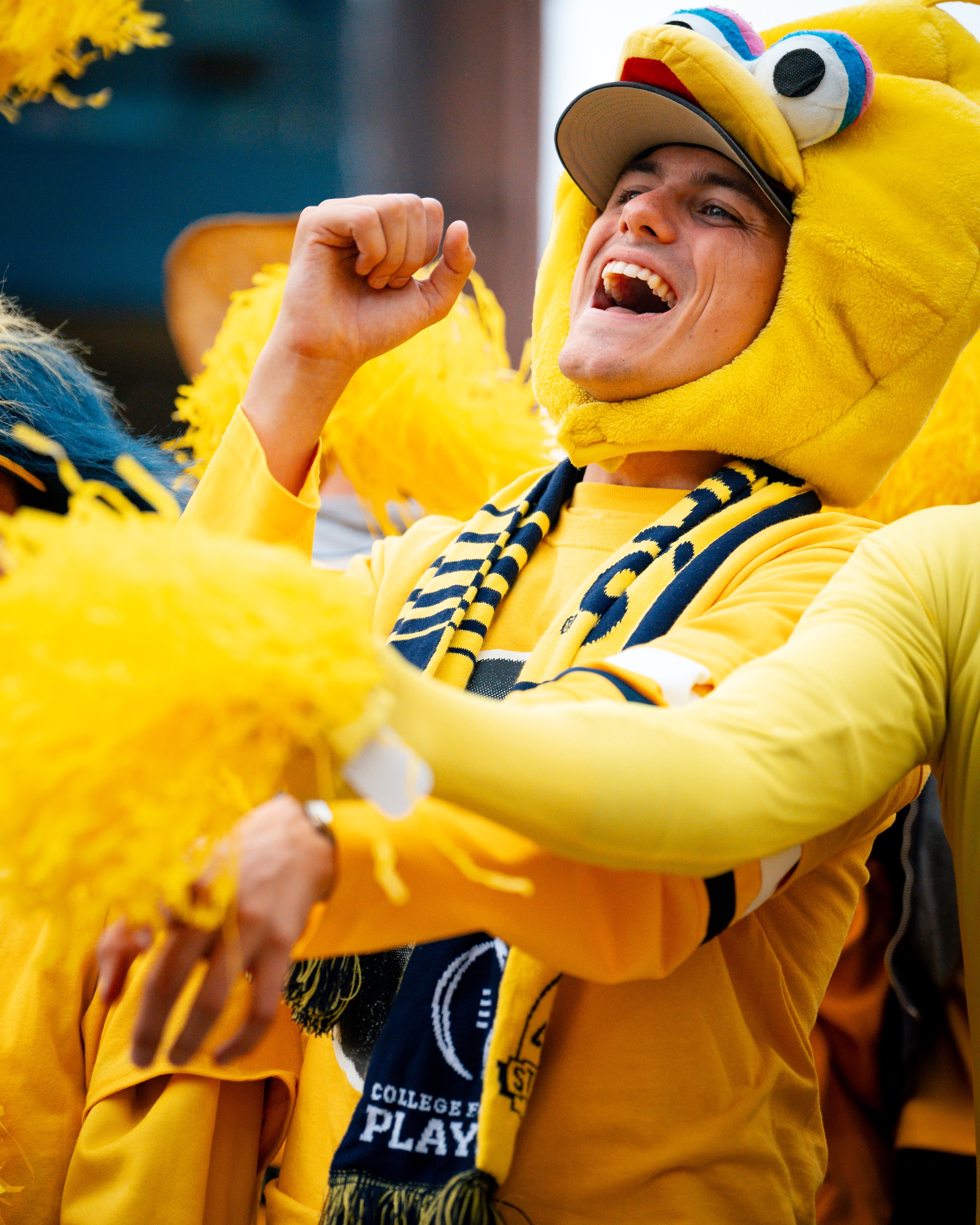 A University of Michigan fan in maize and blue, wearing a yellow mascot head, cheers with yellow pom-poms at a sports event.