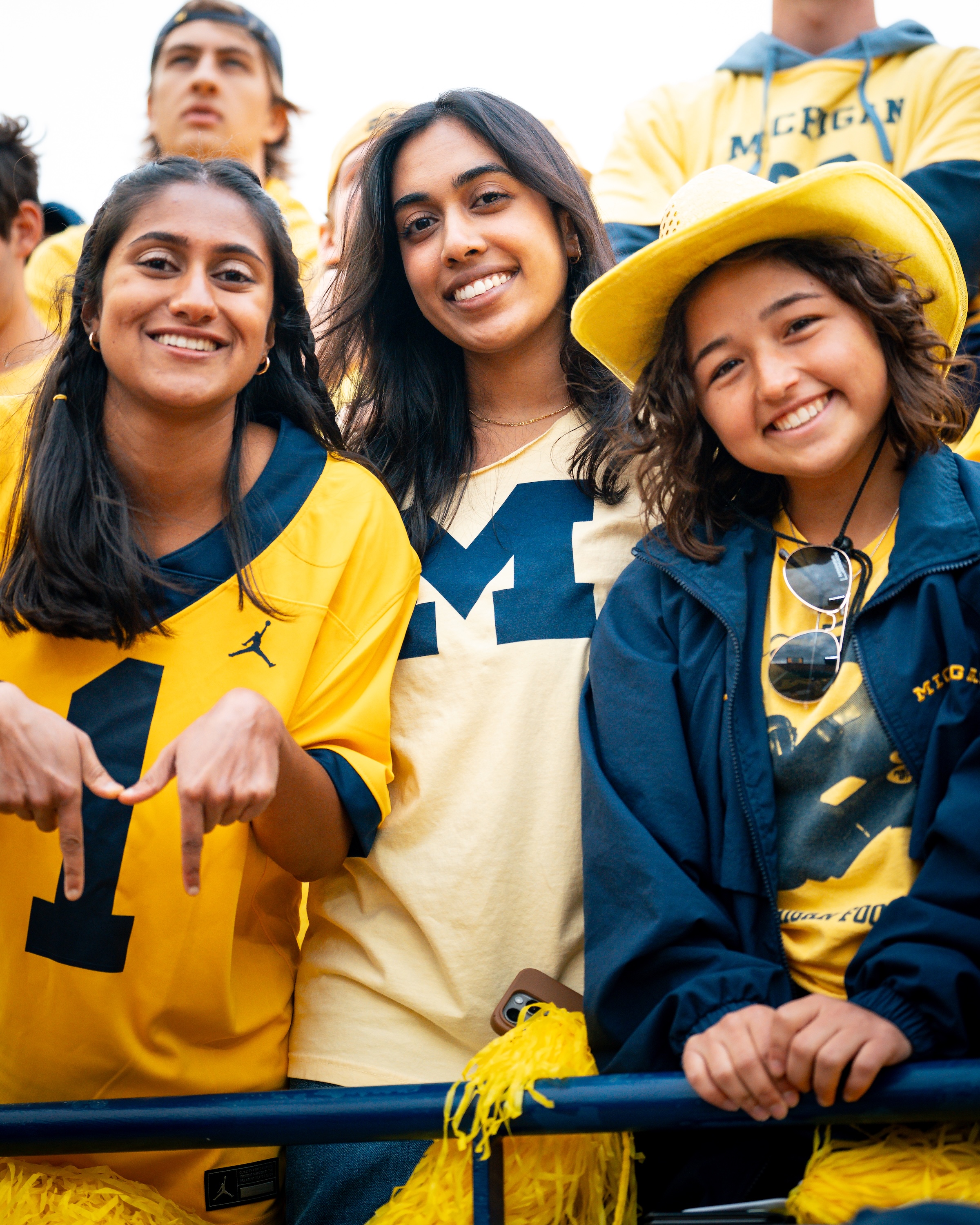 Three smiling young women wearing Michigan apparel in maize and blue at UofM football event. One woman on the left creates a "M" logo with her hands.