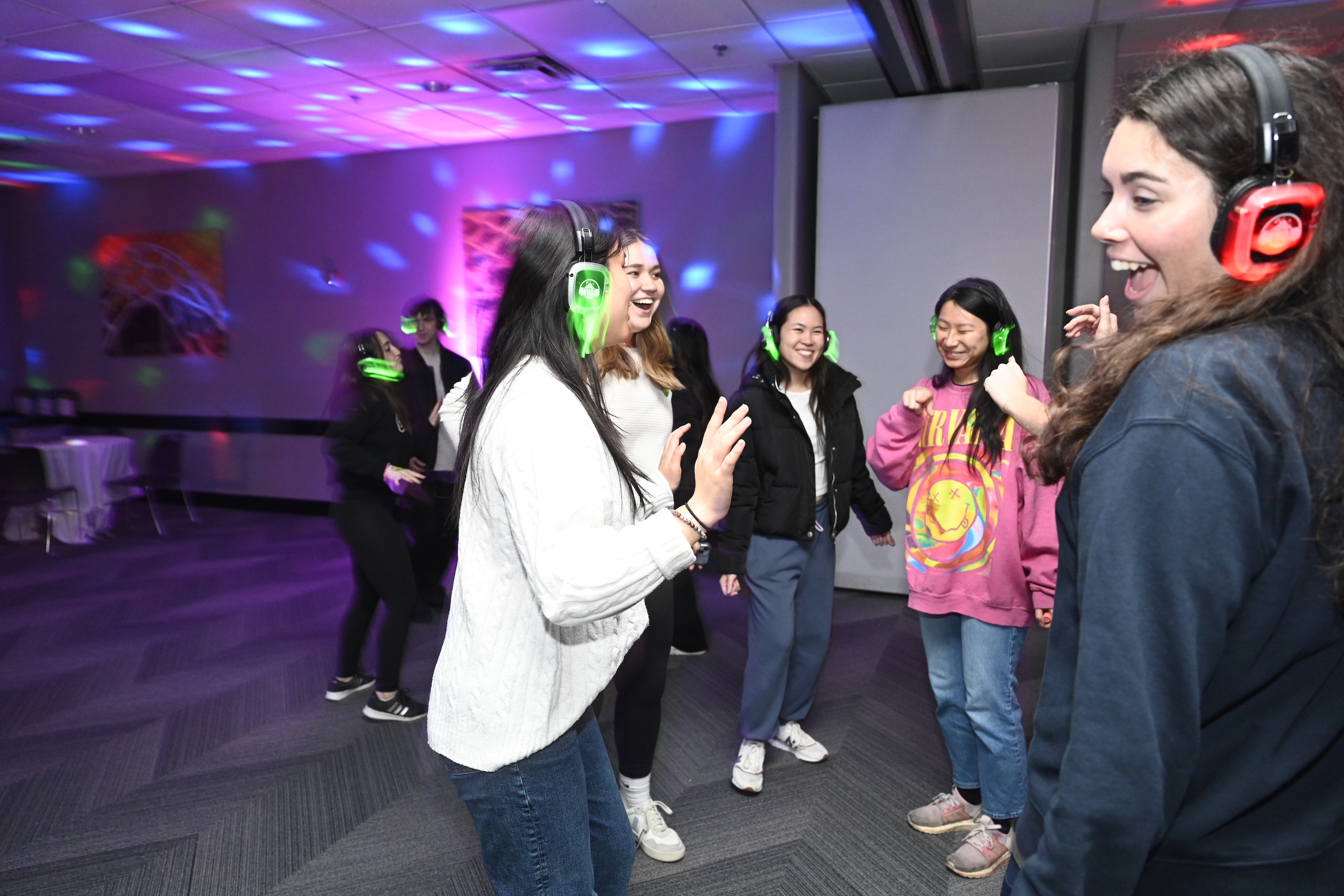 People wearing colorful light-up headphones dance and laugh together at a silent disco party with purple and blue lighting.