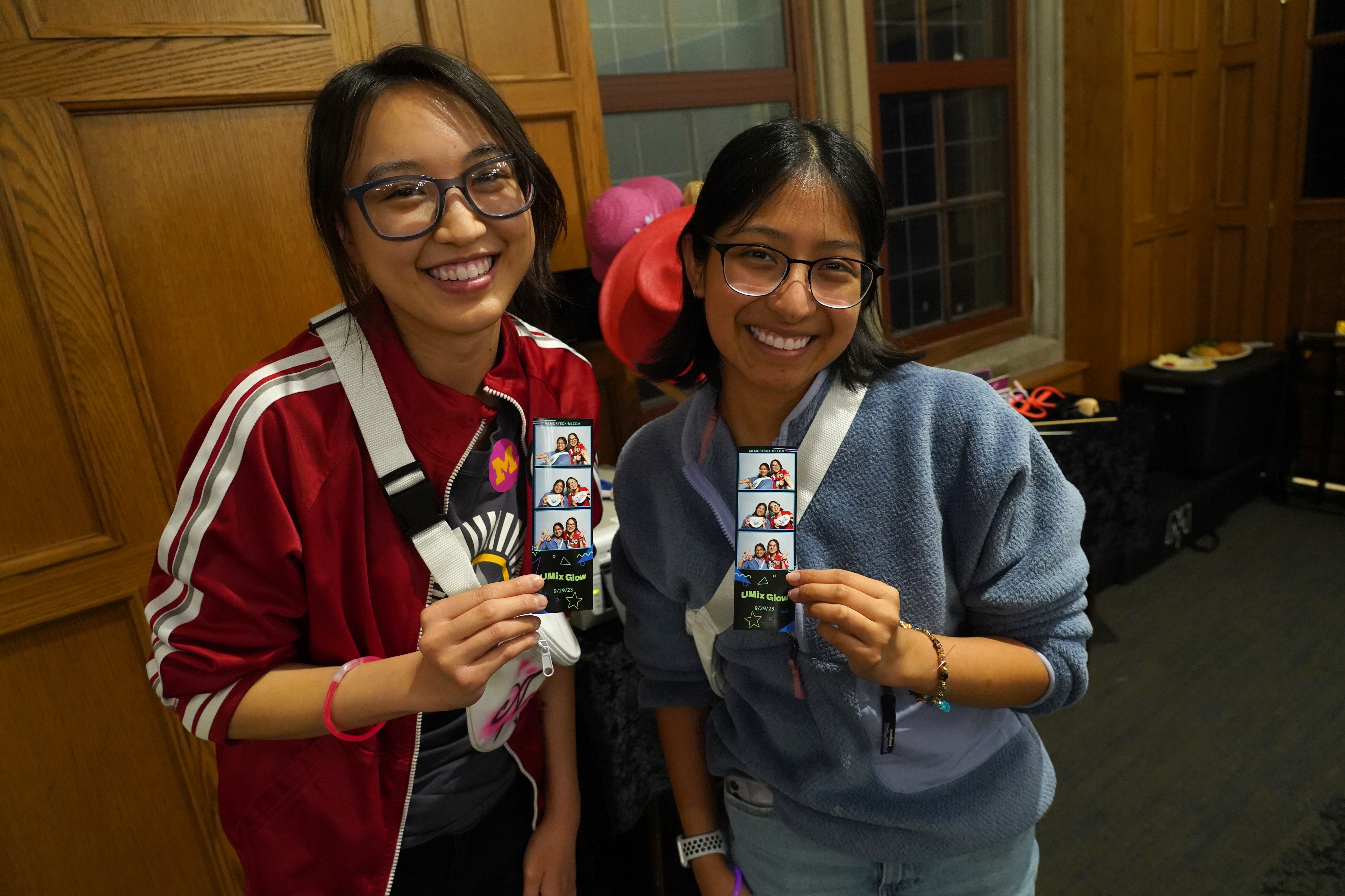 Two students smiling at camera, each holding photo booth strips from UMix Glow event.