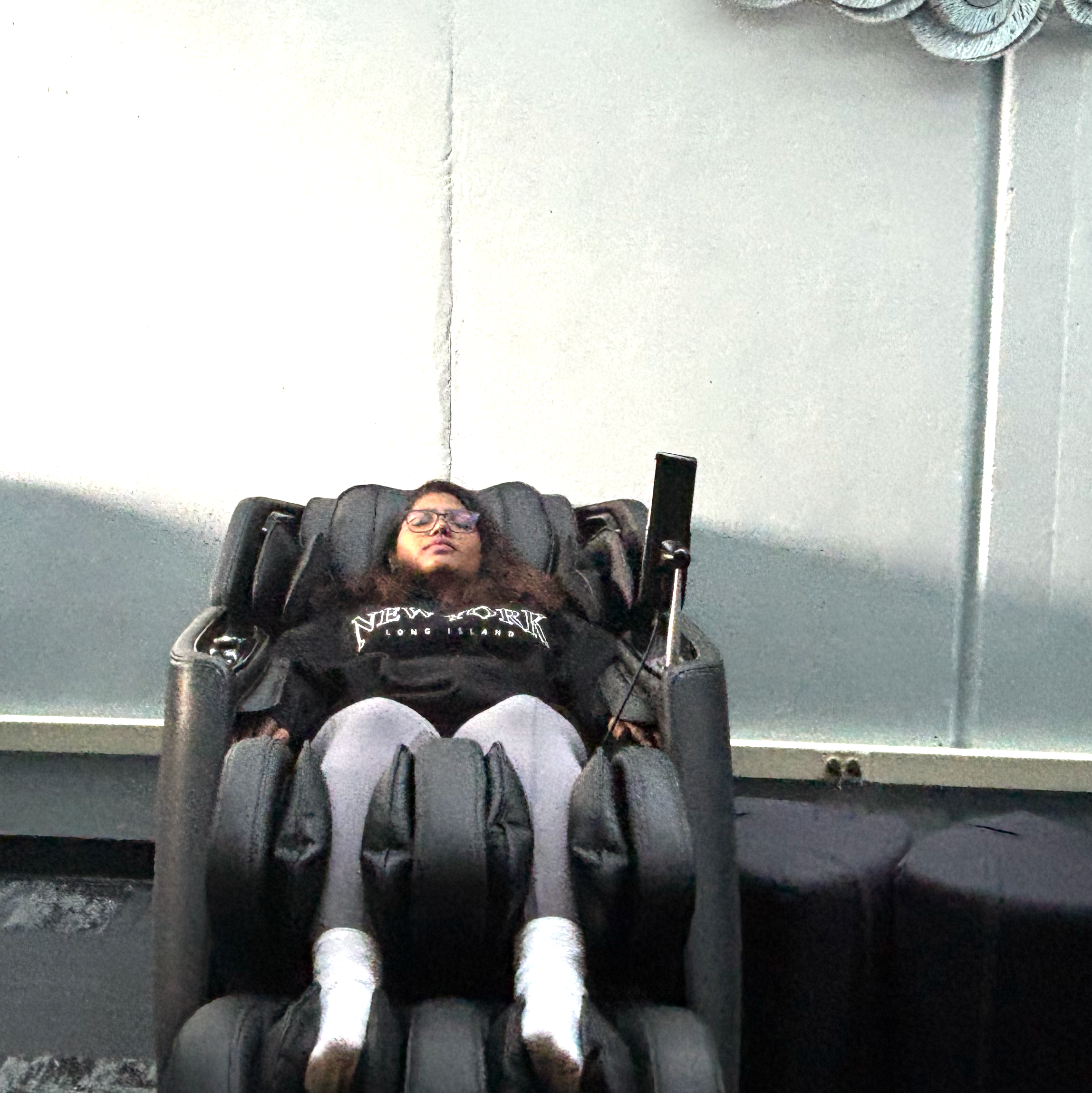 A person lies back in a reclining massage chair with their legs in padded supports, wearing glasses and a black “New York Long Island” sweatshirt.